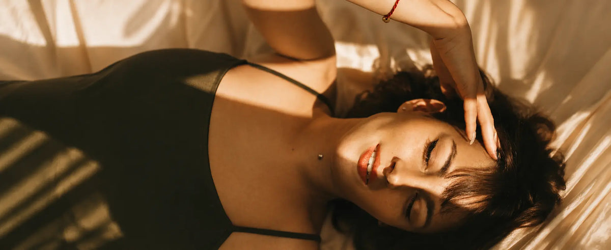 A woman lying down wearing a black camisole, with warm sunlight casting shadows across her face and the fabric background.