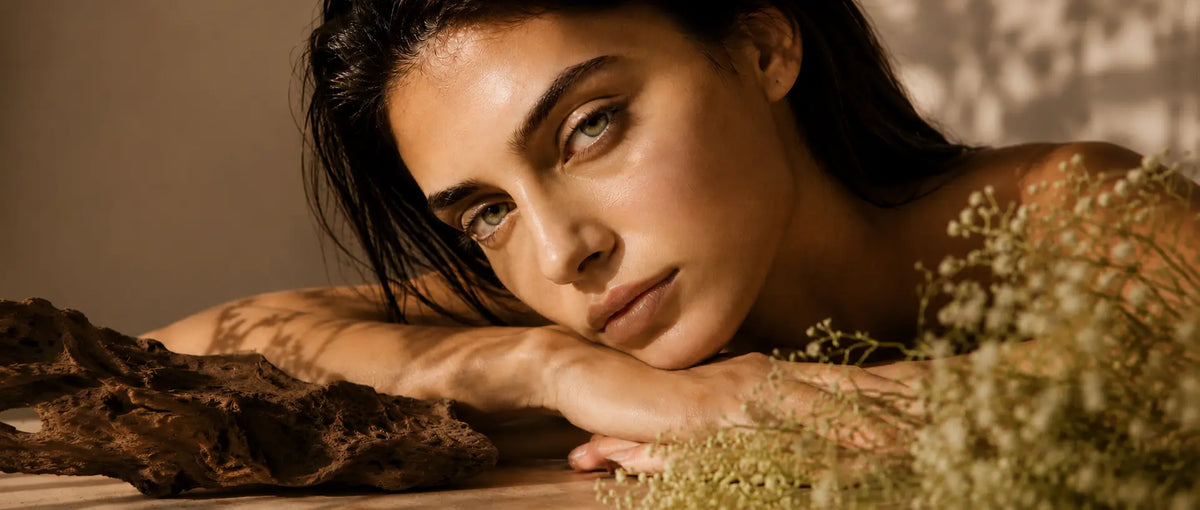 A close-up lifestyle portrait of a woman with dark hair resting her head on her arms, surrounded by dried botanical elements and warm, moody lighting.