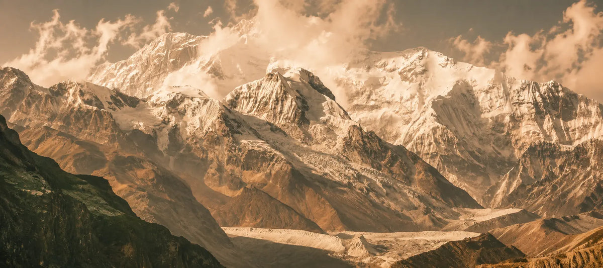 A dramatic mountain landscape with snow-covered peaks, rocky brown slopes, and clouds in warm golden light.