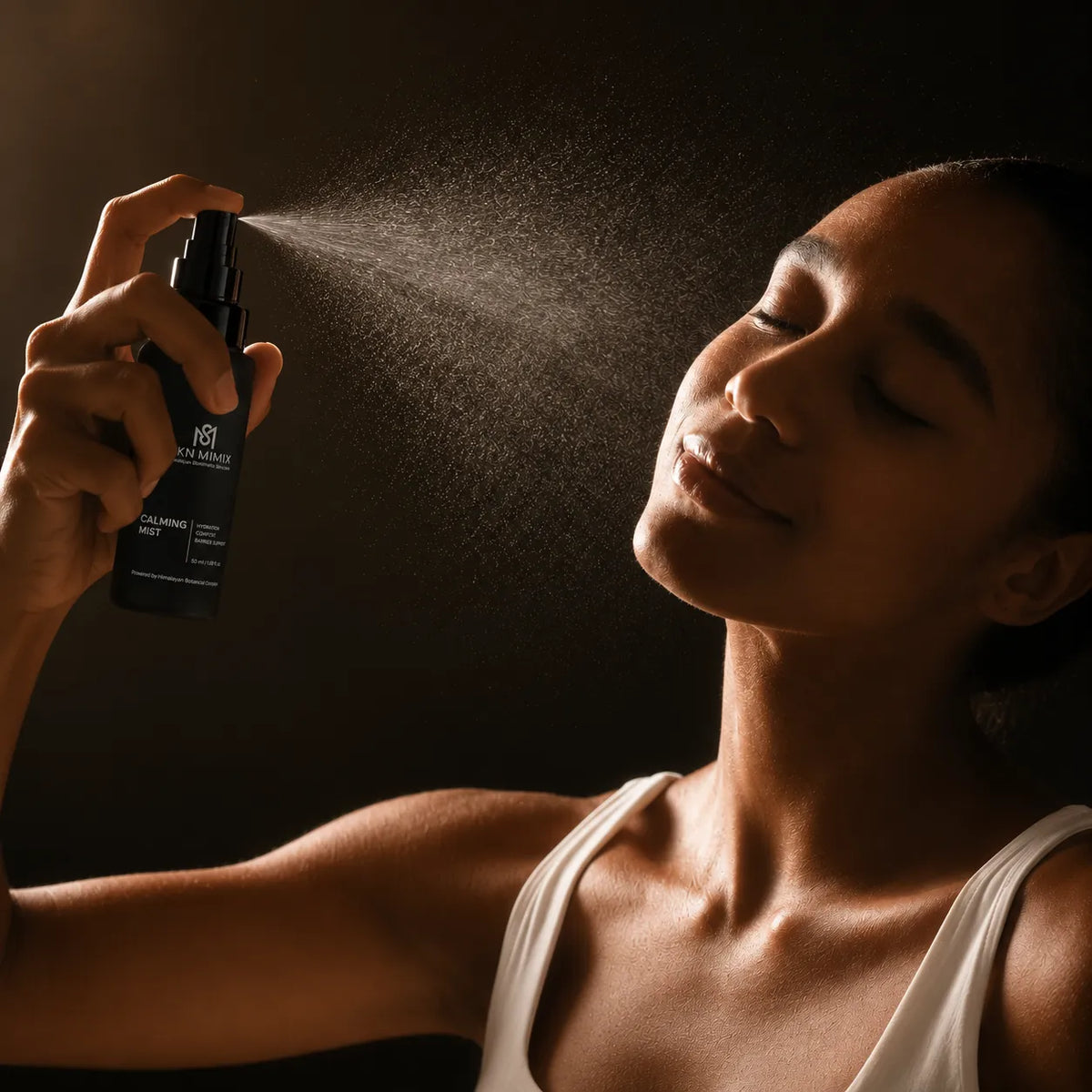 A woman sprays a dark bottle of hair oil onto the air, with the mist visible against a dark background. The bottle has a white logo and text that reads 'H&R' and 'HAIR OIL'.