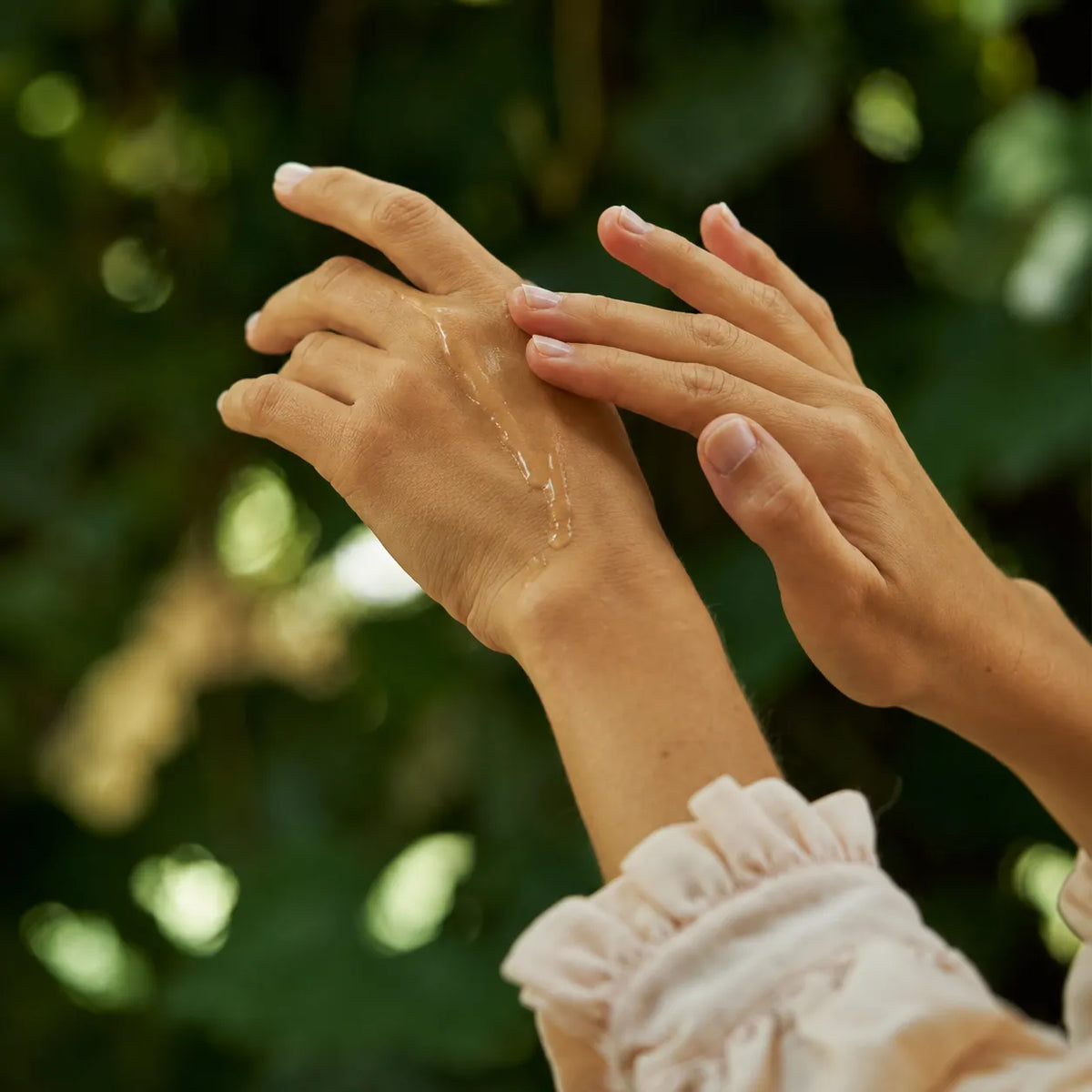 Close-up of a person applying a clear, glossy body oil or serum from a dropper onto the back of their hand, with another hand resting on top, set against a soft, blurred green natural background.